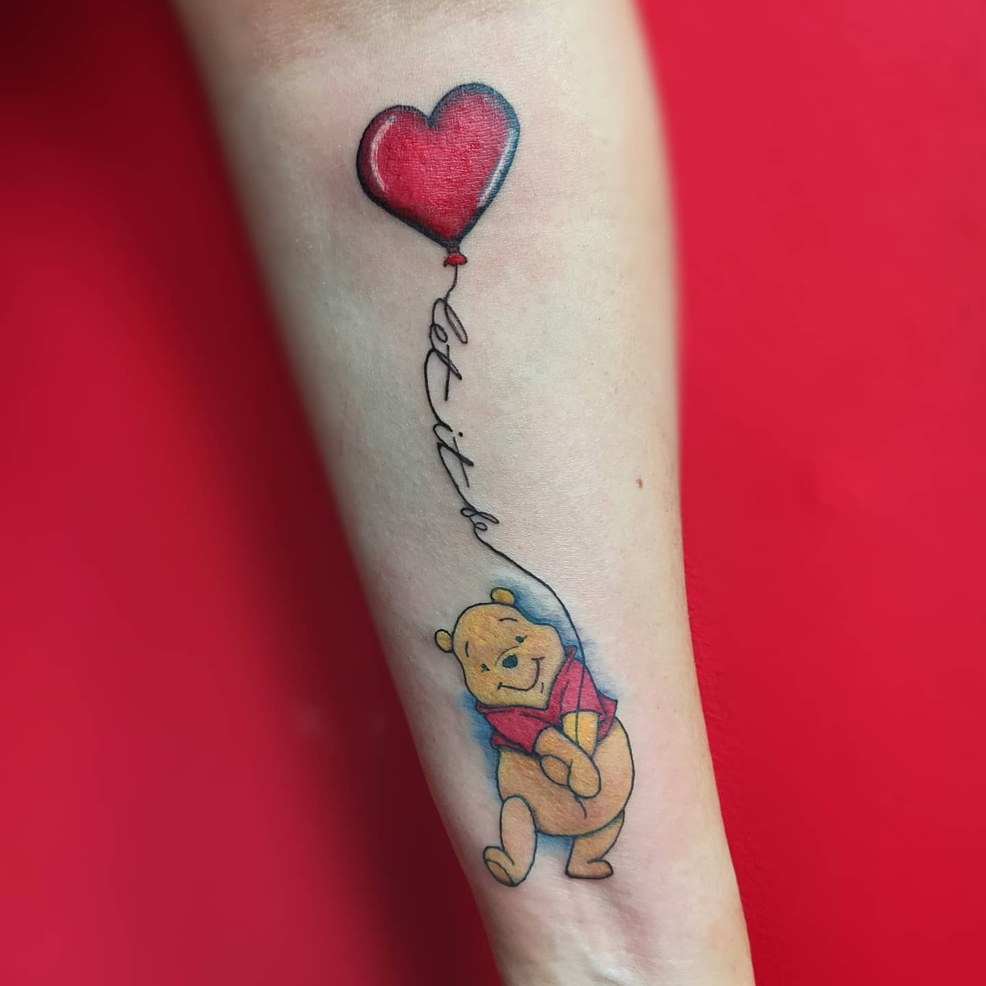 Tattoo of Winnie the Pooh holding a heart balloon with "let it be" text on forearm